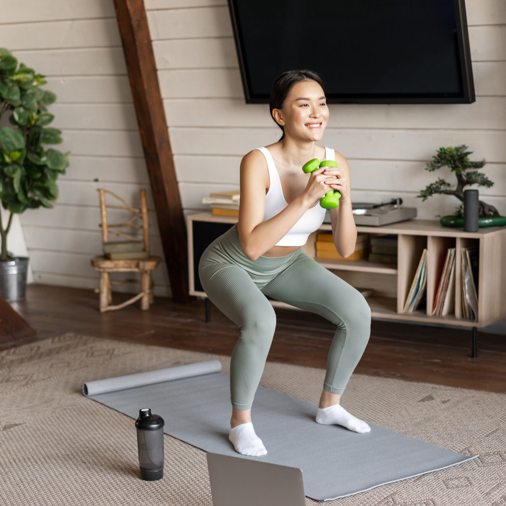Fit and healthy asian woman doing squats at home, workout fitness training session with dumbbells and protein shaker in living room, watching sports video on laptop