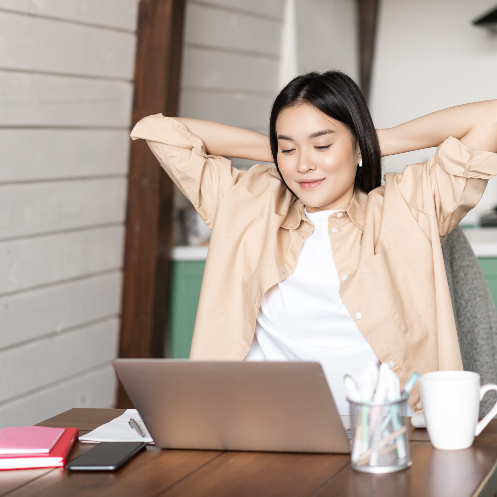 Image of smiling asian girl finish homework or work on laptop, looking pleased at result on computer screen, sitting at kitchen table as her workplace