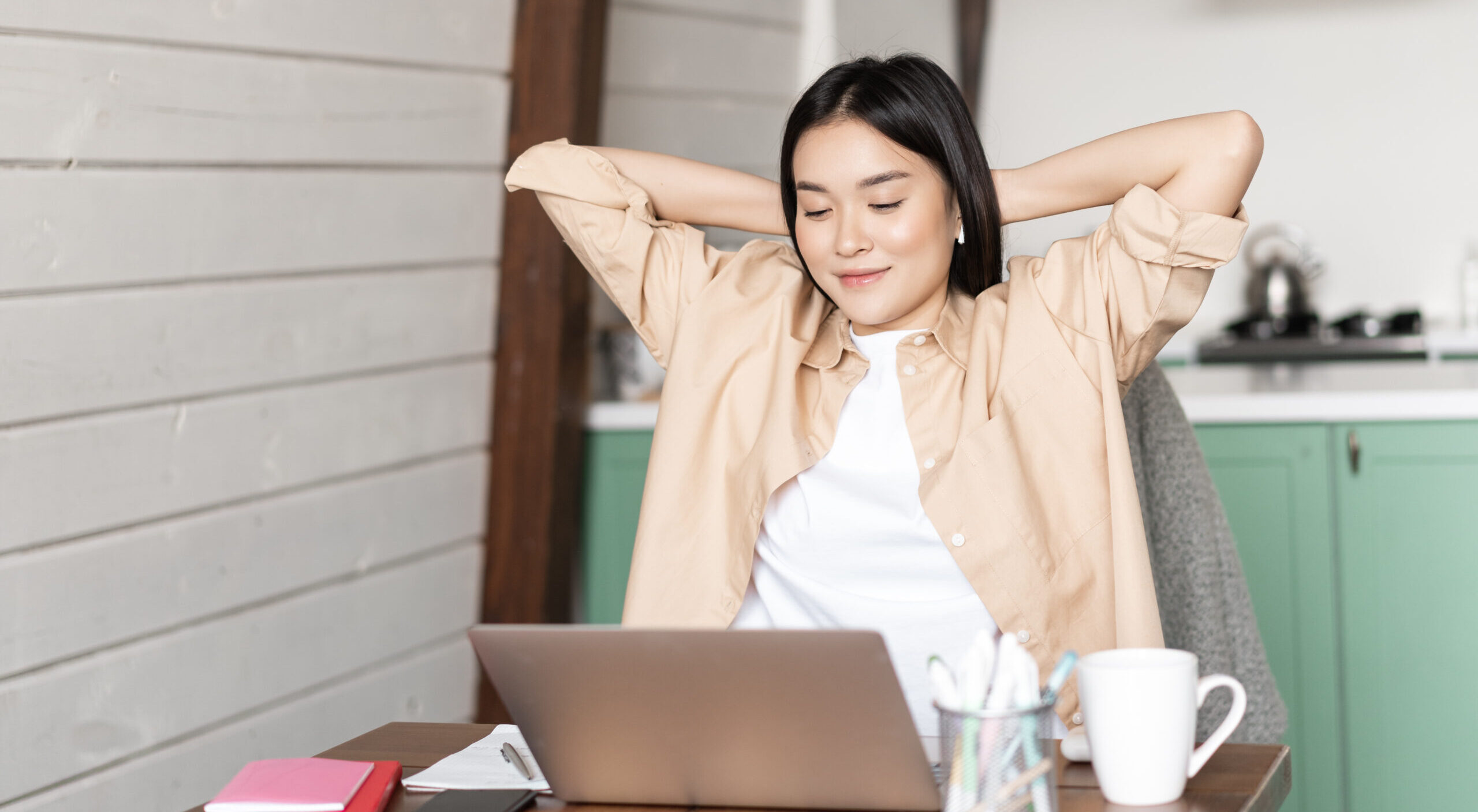 Image of smiling asian girl finish homework or work on laptop, looking pleased at result on computer screen, sitting at kitchen table as her workplace